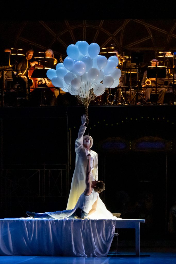 A performer in white holds a bunch of white balloons above another performer lying dramatically on a table, with an orchestra visible in the background.