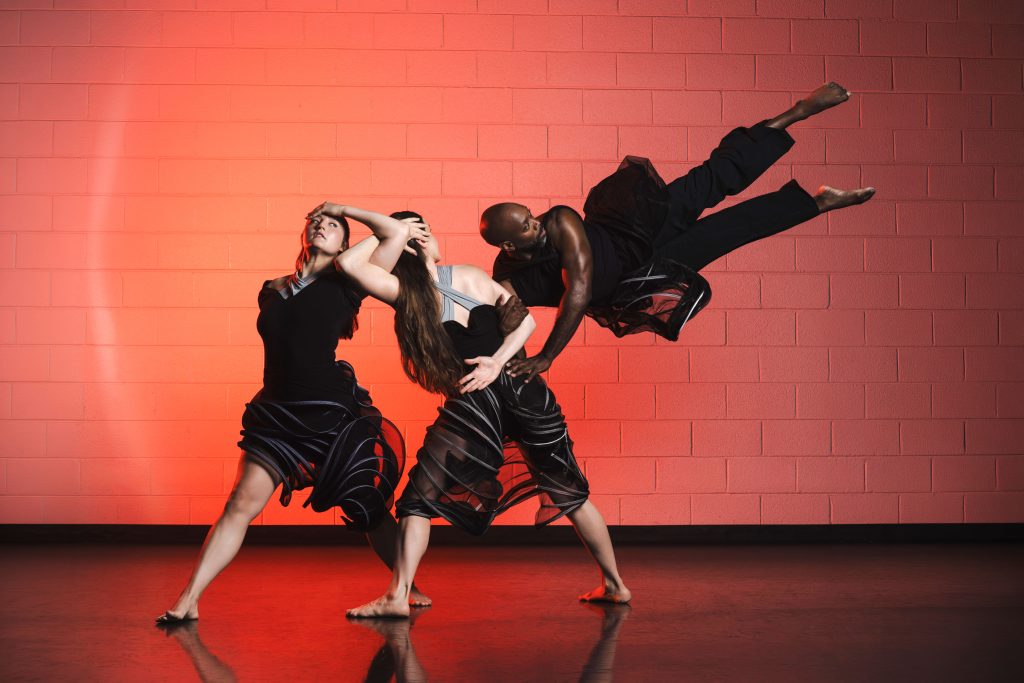 Three barefoot dancers in black outfits perform a dynamic pose against a red brick wall. One dancer is mid-air, supported by the other two, whose arms and bodies create dramatic, fluid lines.