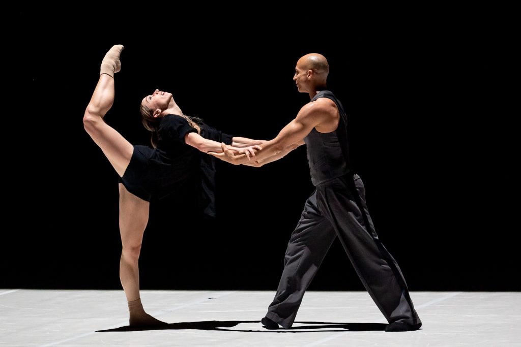 A male and female dancer perform on stage; the woman balances on one leg with her other leg extended high, while the man holds her hands for support.