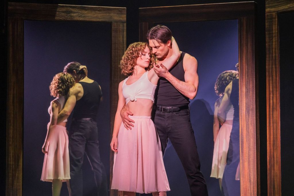 A man and woman in dance attire share an intimate moment on stage, standing in front of large mirrors that reflect their embrace. The lighting is dramatic, highlighting their connection.
