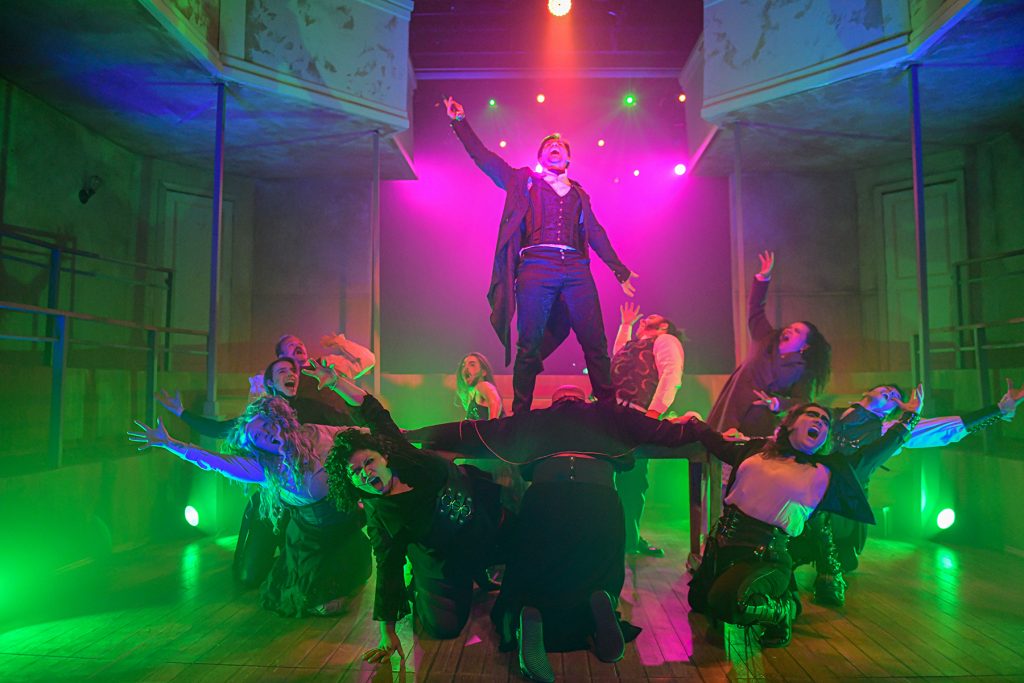 A theatrical performance with a central figure standing triumphantly on a raised platform, surrounded by expressive dancers in dramatic poses, with colorful stage lighting and fog effects.