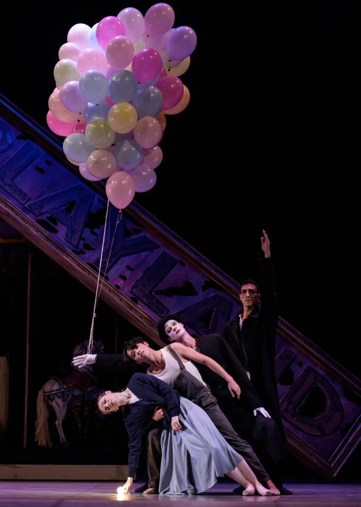 Four dancers pose dramatically on stage, leaning together while holding a bunch of colorful balloons. A large tilted sign and carousel horse are visible in the dimly lit background.