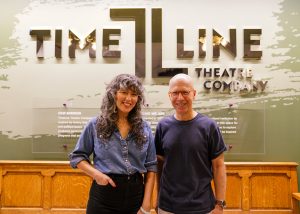 Two people stand smiling in front of a wall sign that reads TimeLine Theatre Company. The person on the left has curly hair and wears a denim shirt; the person on the right has short hair and wears glasses and a dark shirt.