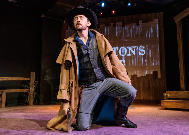 A man dressed as a cowboy kneels on stage holding a revolver, wearing a brown coat and black hat, with a wooden saloon backdrop and dramatic lighting.