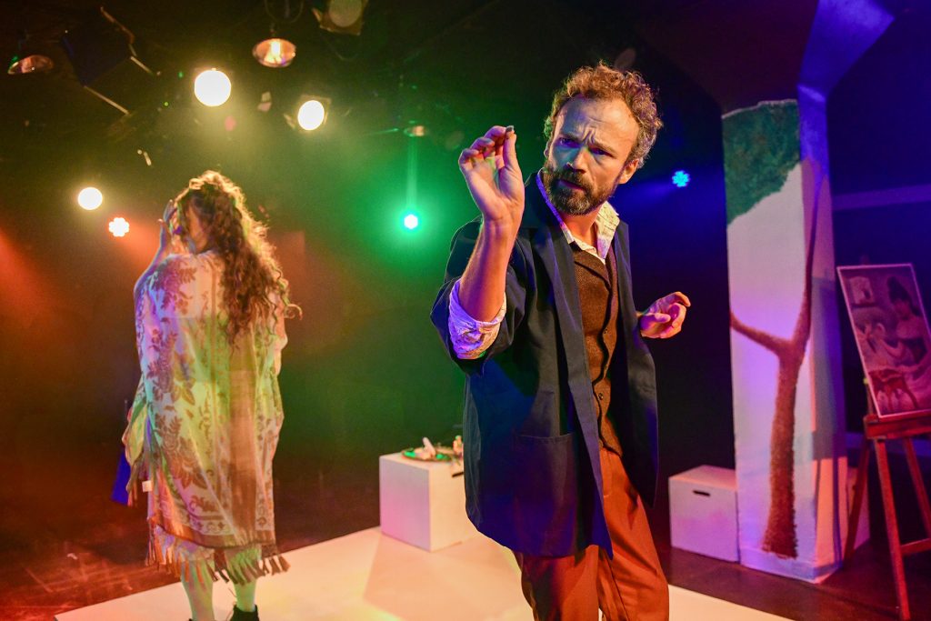 A man gestures dramatically on stage under colorful lights, while a woman with long hair stands behind him. The set includes painted trees and artwork, evoking an artistic or theatrical scene.