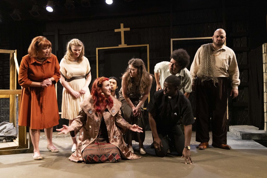 Seven actors on stage, some standing and some kneeling, appear engaged in a dramatic scene. One woman with red hair gestures passionately, while others look at her intently. A cross and simple set pieces are in the background.