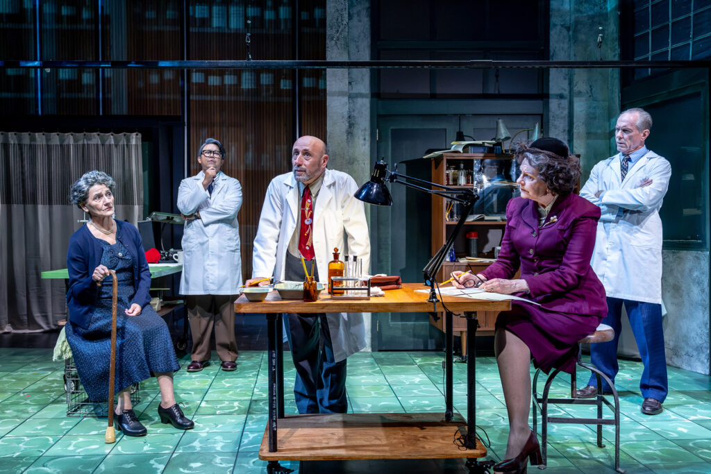 Five actors on a stage set resembling a laboratory; three wear white lab coats, one woman sits with a cane, and another woman sits at a desk in a purple suit, with bottles and lamps on the table.