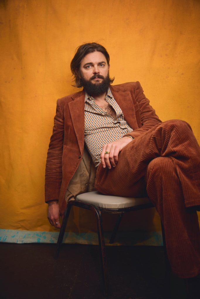 A man with long dark hair and a beard wears a brown corduroy suit and striped shirt, sitting casually on a chair against a mustard yellow background, gazing at the camera.