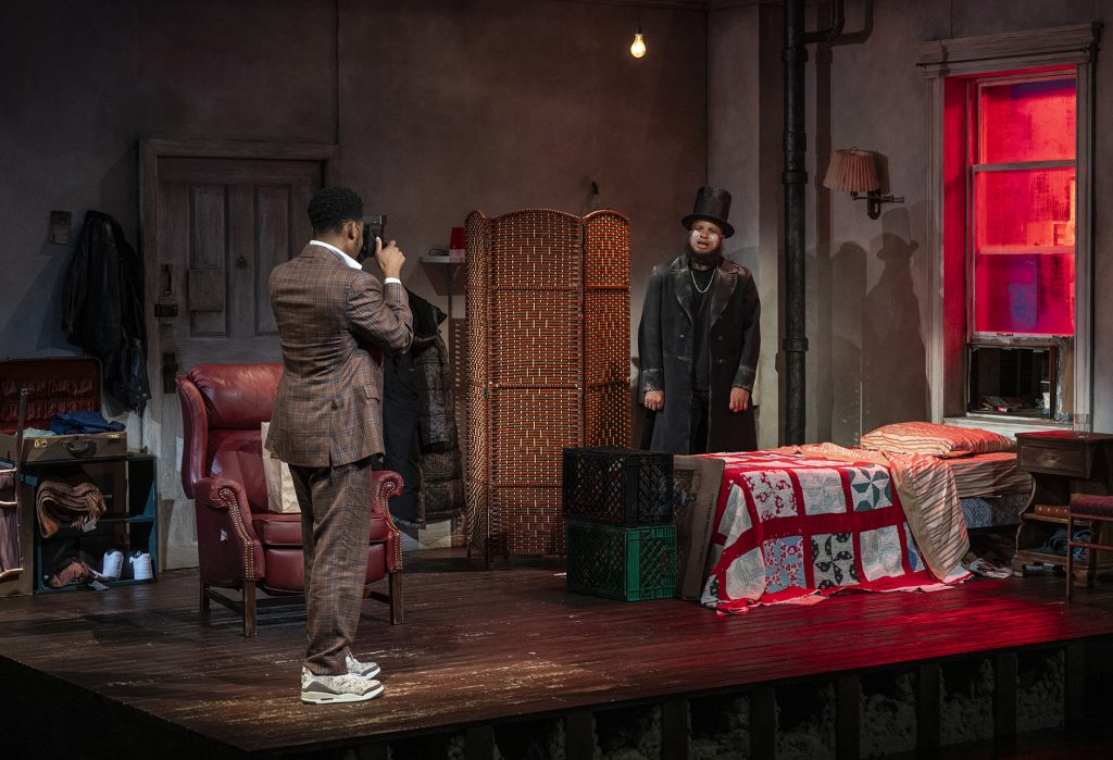 A person in a plaid suit photographs another person wearing a top hat and a long coat inside a dimly lit room with a bed and a red-lit window.